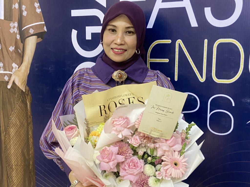 Woman in a purple hijab and striped blouse smiles while holding a large pink flower bouquet and event cards in front of a blue backdrop.