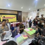 Crowded press conference scene with reporters taking photos and officials seated at a long table in front of a yellow banner that reads 'KONFERENSI PERS'.