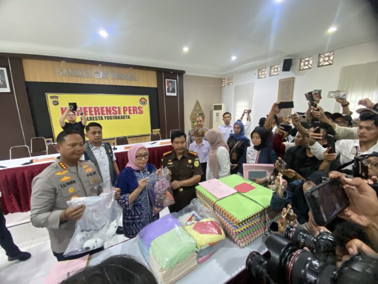 Crowded press conference scene with reporters taking photos and officials seated at a long table in front of a yellow banner that reads 'KONFERENSI PERS'.