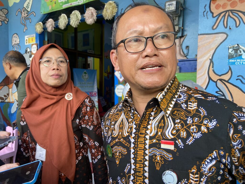 Selfie-style photo of a man in a patterned batik shirt in the foreground and a woman in a brown hijab behind him, against a colorful mural with ocean-themed drawings.