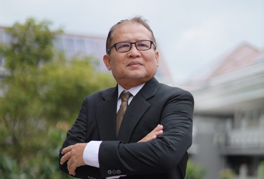 Older man in a dark suit and glasses stands outdoors with arms crossed, looking confident.