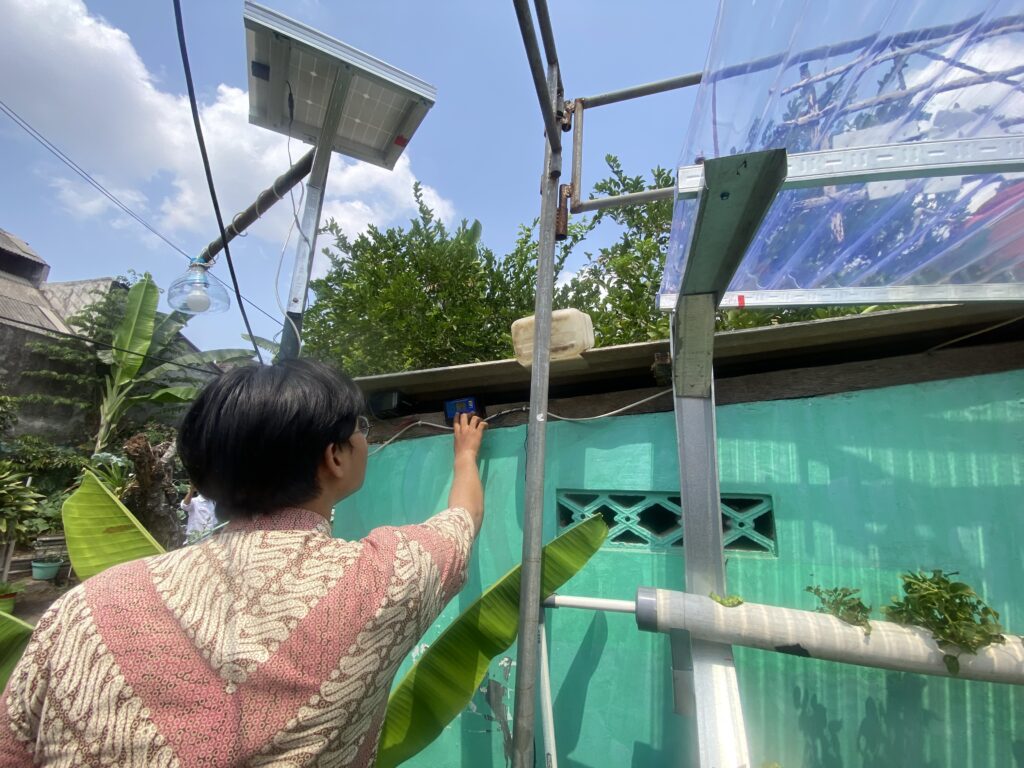 Person in a patterned shirt reaches up to adjust an electrical box on a metal scaffold outdoors, with a solar panel and translucent roofing nearby.