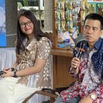 Two people seated at a table outdoors, woman smiling in a beige patterned dress, man in a blue patterned shirt holding a microphone. Snack stall and banner in background.