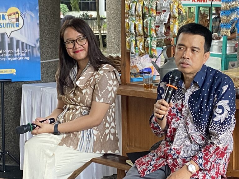 Two people seated at a table outdoors, woman smiling in a beige patterned dress, man in a blue patterned shirt holding a microphone. Snack stall and banner in background.