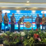 Officials in batik and uniform stand on a stage with a gong centerpiece during an ASEAN cooperative meeting, flags beside them and a blue banner in the background.