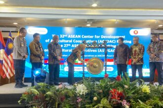 Officials in batik and uniform stand on a stage with a gong centerpiece during an ASEAN cooperative meeting, flags beside them and a blue banner in the background.