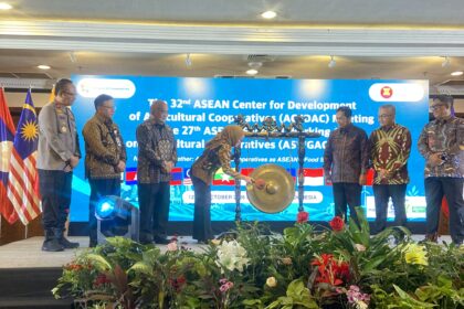Officials in batik and uniform stand on a stage with a gong centerpiece during an ASEAN cooperative meeting, flags beside them and a blue banner in the background.
