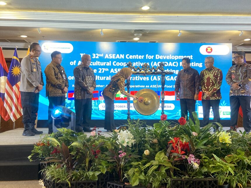 Officials in batik and uniform stand on a stage with a gong centerpiece during an ASEAN cooperative meeting, flags beside them and a blue banner in the background.