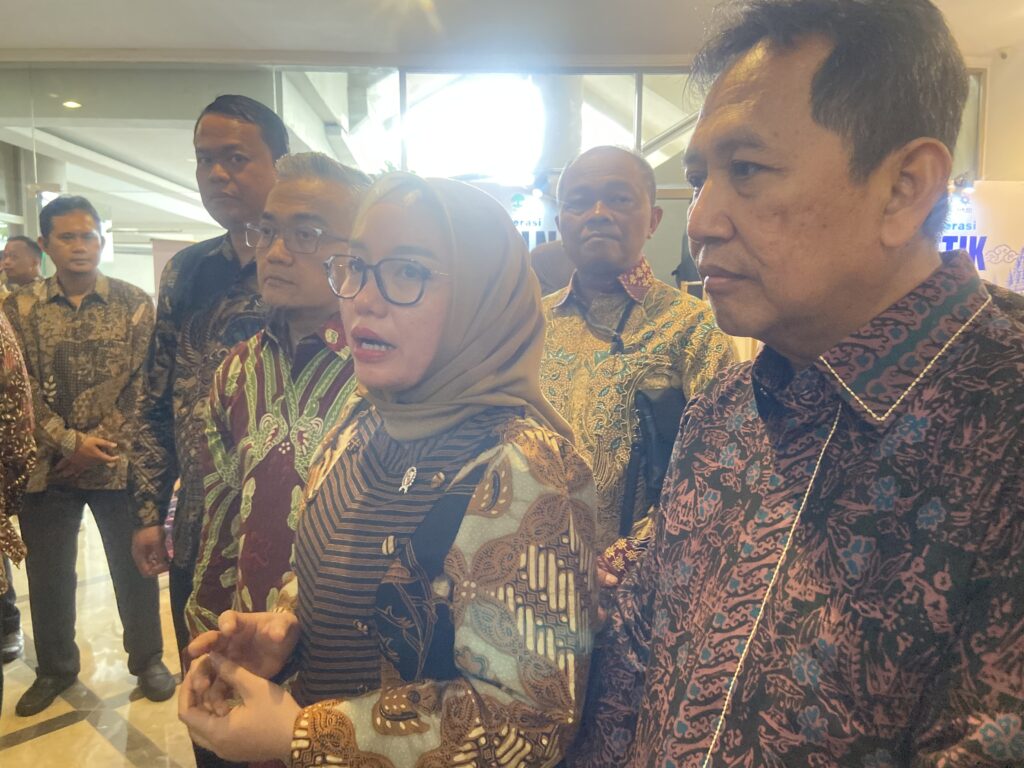 IMG_5635 - Mabur.co Woman in a hijab and glasses in the foreground, flanked by men in batik shirts at an indoor event.