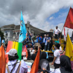 Crowd at a street protest with colorful flags and a speaker on a truck stage near a building near a blue sky.