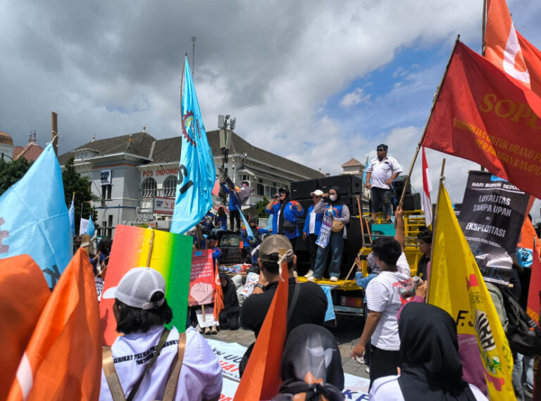 Crowd at a street protest with colorful flags and a speaker on a truck stage near a building near a blue sky.