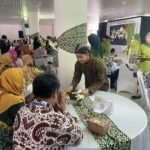 People sit at round tables during a cultural banquet; attendees wear batik and hijabs, with a server in brown batik moving among them.