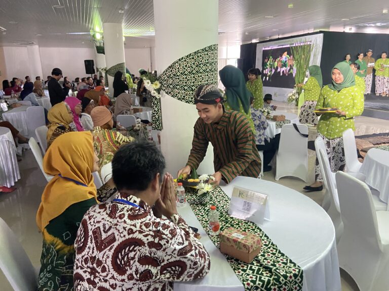 People sit at round tables during a cultural banquet; attendees wear batik and hijabs, with a server in brown batik moving among them.