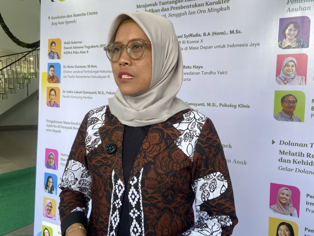 Woman wearing a light gray hijab and round glasses at a conference, standing in front of a banner with speakers’ photos and text behind her.