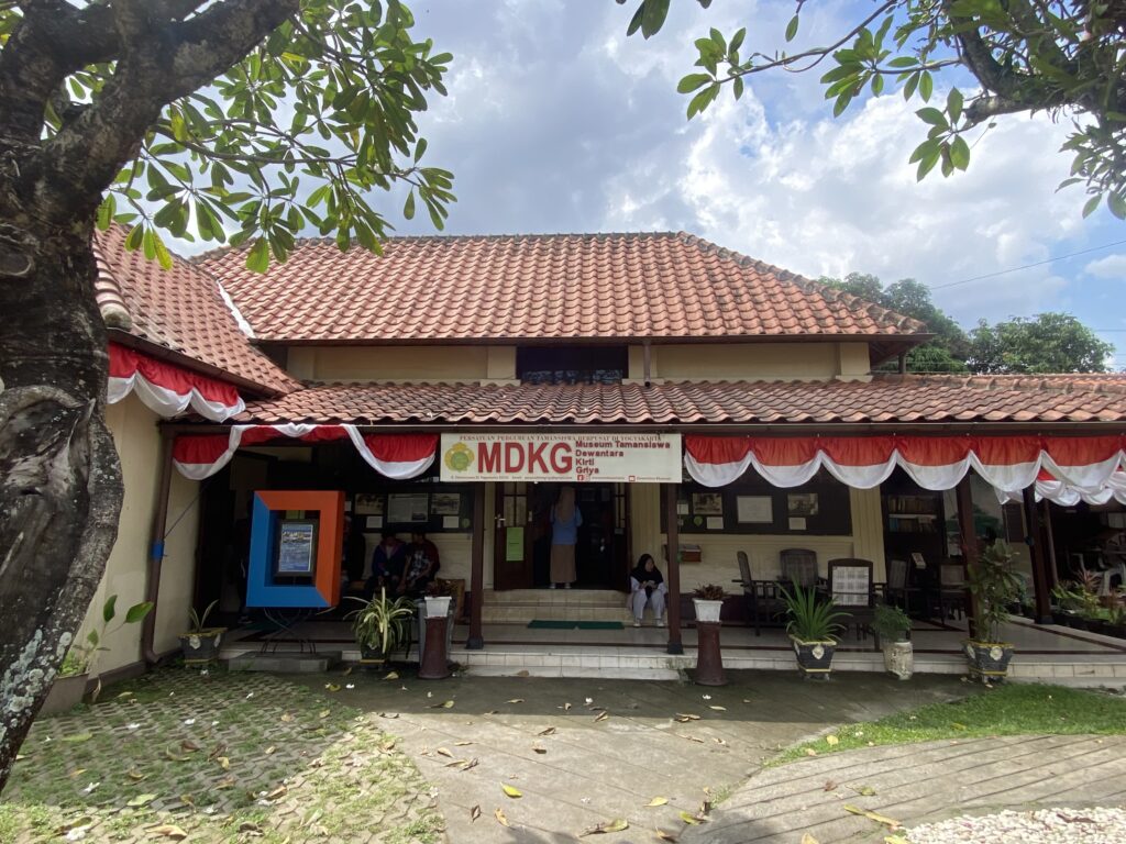 Front view of a beige building with a red-tiled roof, red-and-white bunting along the eaves, and a banner reading 'MDKG' (Museum Tamansiswa) on the entrance.
