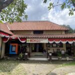Front view of a beige building with a red-tiled roof, red-and-white bunting along the eaves, and a banner reading 'MDKG' (Museum Tamansiswa) on the entrance.