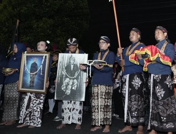 IMG_5678 - Mabur.co A nighttime street procession with people in traditional batik clothing; some carry framed black-and-white portraits draped with garlands while others hold ceremonial staffs and ceremonial garments nearby.