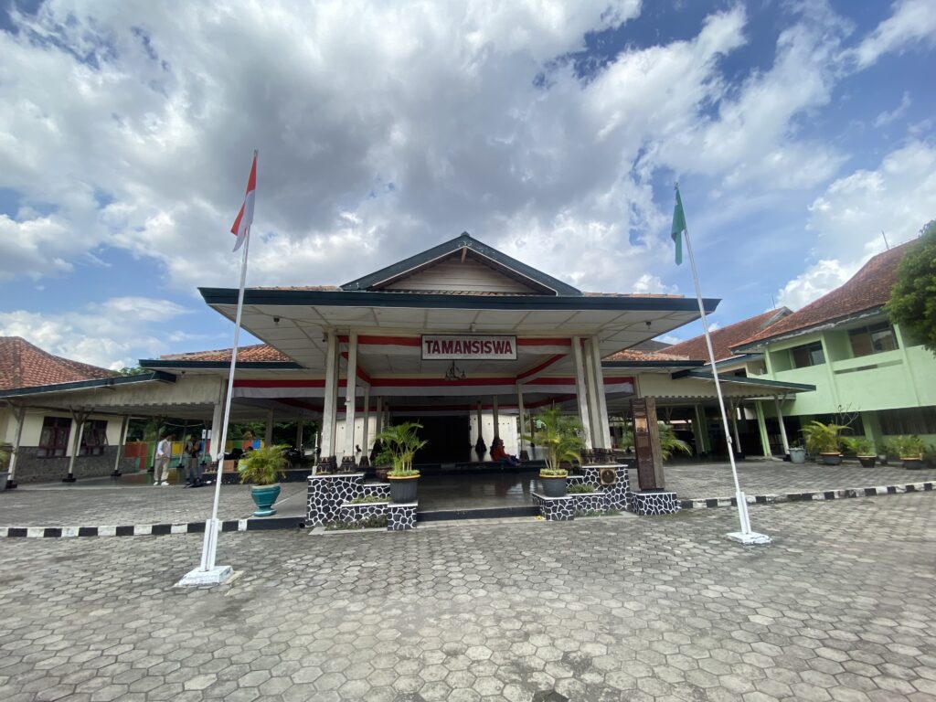 Open-air school courtyard with a raised stage, red-and-white and green flags on tall poles, and potted plants.