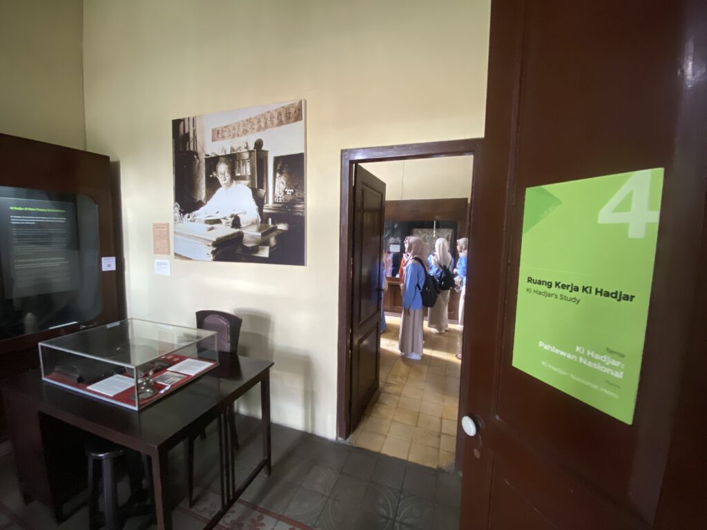 Exhibit room with a large black-and-white portrait of a man at a desk on the wall and glass display case in the foreground. A doorway shows visitors in blue uniforms beyond the door, and a green poster is mounted on the door.
