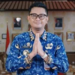 Man in a blue batik shirt with hands pressed together in a respectful greeting gesture, indoors.