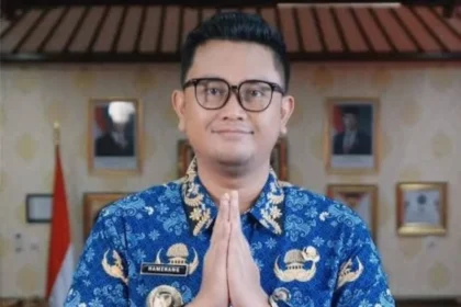 Man in a blue batik shirt with hands pressed together in a respectful greeting gesture, indoors.