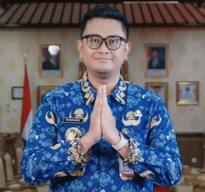 Man in a blue batik shirt with hands pressed together in a respectful greeting gesture, indoors.