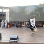 Outdoor stage with a large LED screen, two masked performers in costumes, and stage gear in front.
