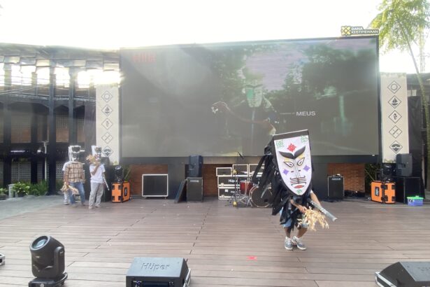 Outdoor stage with a large LED screen, two masked performers in costumes, and stage gear in front.