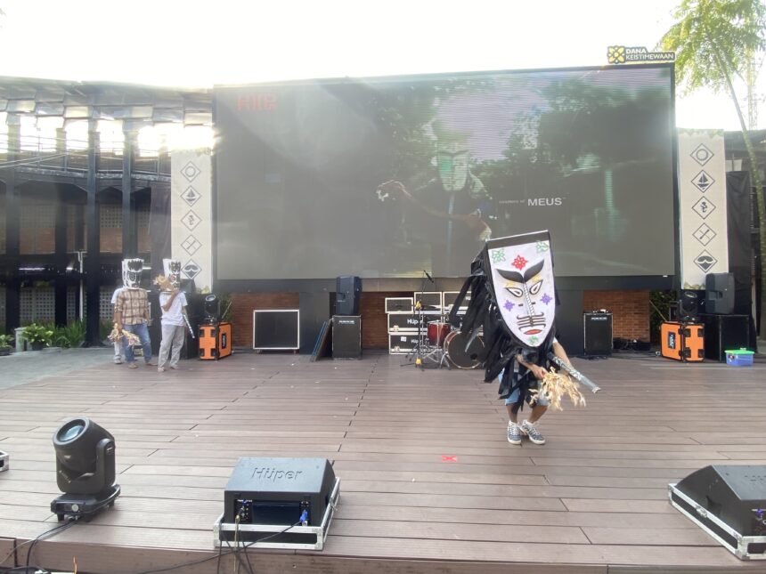 Outdoor stage with a large LED screen, two masked performers in costumes, and stage gear in front.