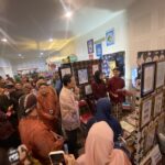 Crowded indoor craft fair with people browsing textile displays and framed artwork along the walls.