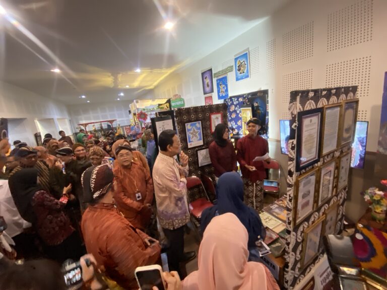 Crowded indoor craft fair with people browsing textile displays and framed artwork along the walls.