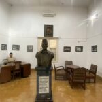 Bronze bust on a pedestal in a museum-like room, with framed black-and-white photos on white walls and wooden chairs around.