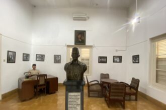 Bronze bust on a pedestal in a museum-like room, with framed black-and-white photos on white walls and wooden chairs around.