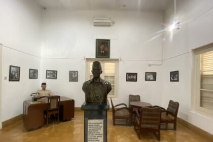 Bronze bust on a pedestal in a museum-like room, with framed black-and-white photos on white walls and wooden chairs around.