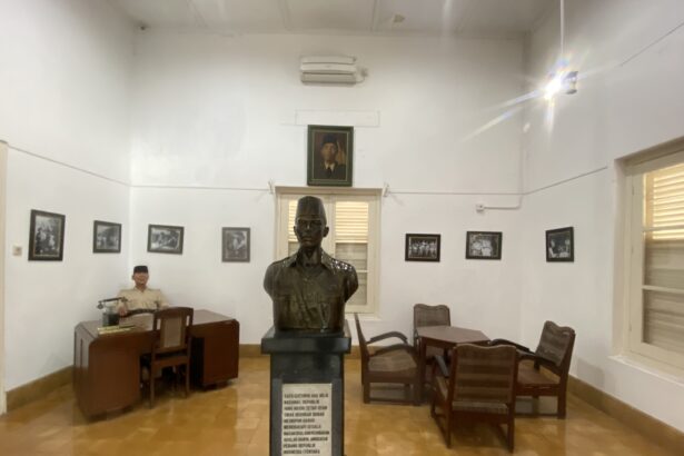 Bronze bust on a pedestal in a museum-like room, with framed black-and-white photos on white walls and wooden chairs around.