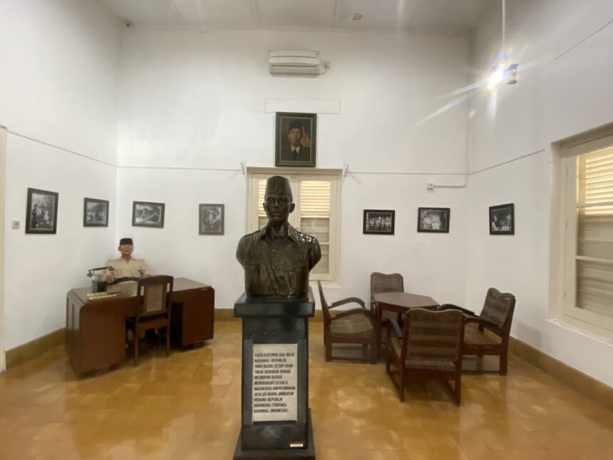Bronze bust on a pedestal in a museum-like room, with framed black-and-white photos on white walls and wooden chairs around.