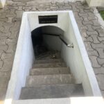 Open concrete stairwell downward with a low ceiling and a sign that reads Keep Your Head / Harap Kepala Menunduk, warning to duck as you descend