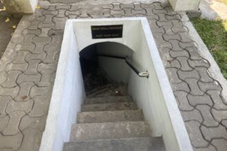 Open concrete stairwell downward with a low ceiling and a sign that reads Keep Your Head / Harap Kepala Menunduk, warning to duck as you descend