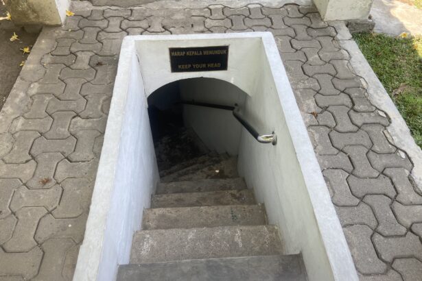 Open concrete stairwell downward with a low ceiling and a sign that reads Keep Your Head / Harap Kepala Menunduk, warning to duck as you descend