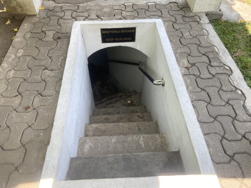 Open concrete stairwell downward with a low ceiling and a sign that reads Keep Your Head / Harap Kepala Menunduk, warning to duck as you descend