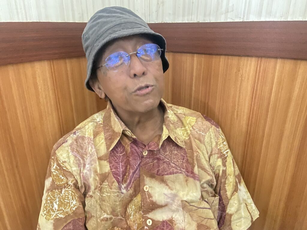 Older man in a gray bucket hat and blue-reflection glasses, wearing a multicolor batik shirt, seated against a wooden panel wall.