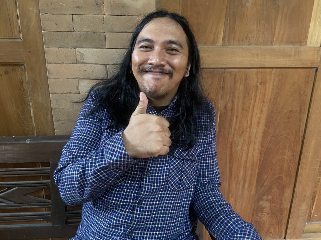 Man with long black hair and a mustache, wearing a blue plaid shirt, giving a thumbs up indoors.
