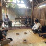 Group of people seated on the floor around a central snack tray inside a rustic wooden pavilion, engaged in a casual meeting.