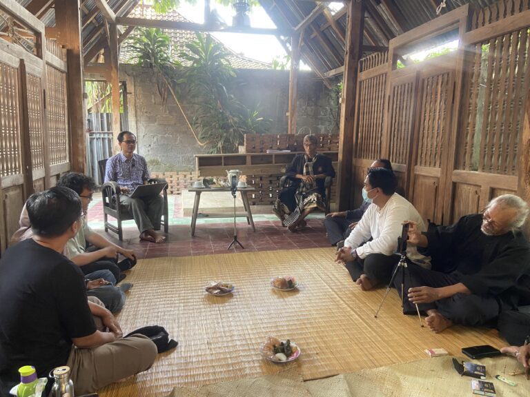 Group of people seated on the floor around a central snack tray inside a rustic wooden pavilion, engaged in a casual meeting.