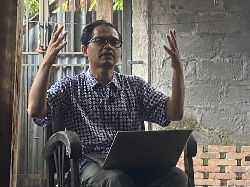 Man wearing a patterned shirt and glasses raises both hands while seated at a desk with a laptop, in a rustic outdoor setting.