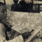 Woman sits at a quilt, sewing intricate patterns with a needle in hand (black-and-white photo).