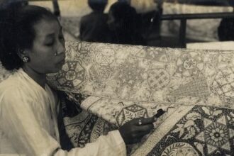 Woman sits at a quilt, sewing intricate patterns with a needle in hand (black-and-white photo).