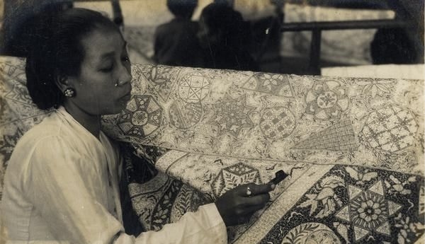 Woman sits at a quilt, sewing intricate patterns with a needle in hand (black-and-white photo).
