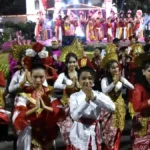 Group of people in colorful traditional costumes perform a dance at a night festival, hands pressed together in a greeting pose. A stage with performers is in the background.