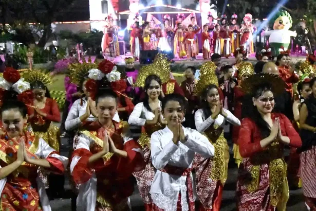Group of people in colorful traditional costumes perform a dance at a night festival, hands pressed together in a greeting pose. A stage with performers is in the background.